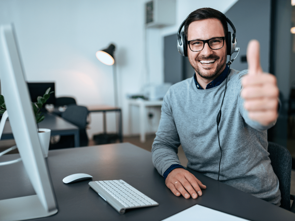 Happy man in grey jumper siting smiling on the right of the image wearing glasses, a headset and holding his thumb up